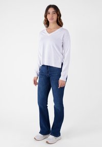 Maglione bianco a maniche lunghe con scollatura a V e orlo a coste, abbinato a jeans flare blu. Le sneakers bianche con suola testurizzata completano l'outfit.