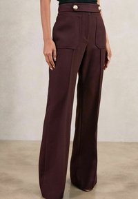 Egy nő magas derekú, széles szárú, sötétlila nadrágot visel, elülső zsebekkel és díszes arany gombokkal a derékpántnál.