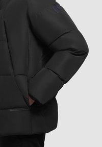 Sort puffer-jakke med en lynlåslomme og stikninger. Har en logo-patch på skulderen og en quiltet tekstur.