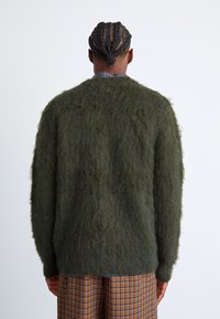 Πράσινο αφράτο πουλόβερ με άνετη εφαρμογή και υφή, φορεμένο πάνω από καφέ καρό παντελόνια. Το πουλόβερ έχει στρογγυλή λαιμόκοψη και μακριά μανίκια.