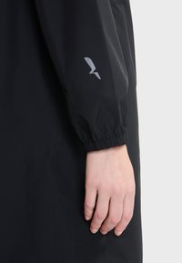 Manica di giacca impermeabile nera con polsino elastico, caratterizzata da un dettaglio del logo in argento. La texture liscia e il design aderente sono caratteristiche notevoli.