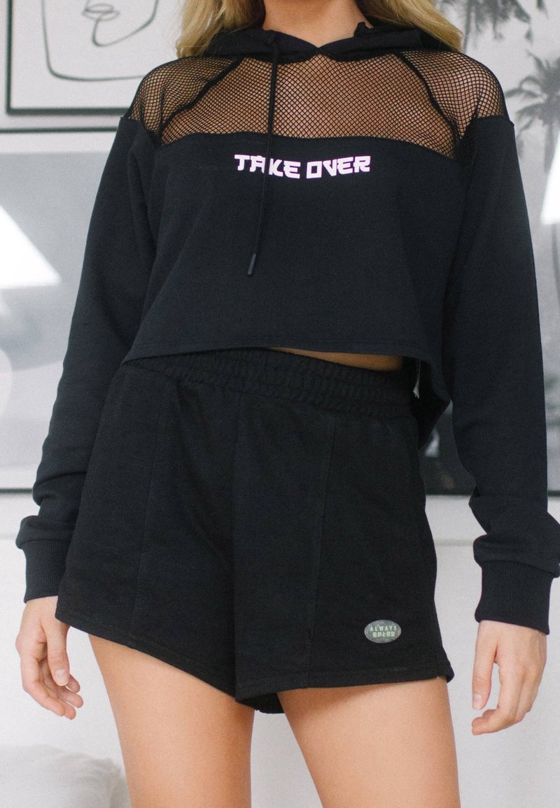 Femme portant un sweat à capuche court noir avec un haut en mesh et le texte "TAKE OVER", associé à un short noir à taille élastique avec une petite étiquette logo.