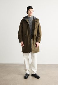 Parka impermeável verde-oliva com capuz, cintura ajustável com cordão e forro cinza, combinado com calças brancas e sapatos pretos sem atacadores.