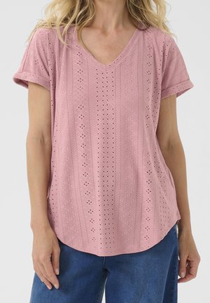 Kobieta ubrana w różową bluzkę z krótkim rękawem o ażurowym wzorze, z dekoltem w serek i podwiniętymi mankietami, w połączeniu z niebieskimi dżinsami.