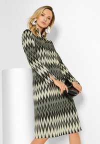Frau in einem knielangen, langärmeligen Kleid mit Zickzackmuster, die eine schwarze Clutch hält und große weiße abstrakte Ohrringe trägt.