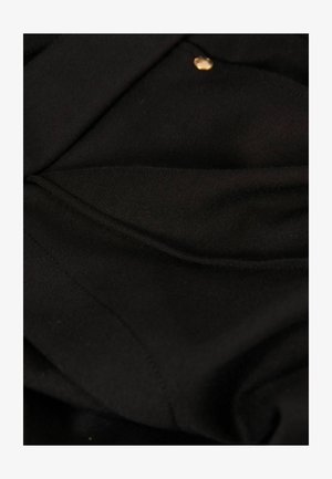 Zwarte stof met een gladde textuur, voorzien van een gouden knop en zichtbare naden, wat duidt op een eenvoudig, gestructureerd ontwerp.