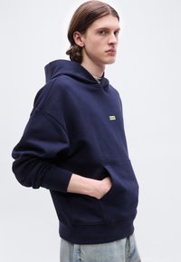 Νεαρός με μακριά μαλλιά που φοράει μπλε σκούρα φούτερ με κουκούλα και μια μικρή κίτρινη ετικέτα «Hugo», έχοντας τα χέρια στην μπροστινή τσέπη, με ανοιχτόχρωμο τζιν.