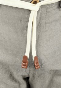 Tissu gris clair avec un motif texturé, cordon blanc avec des accents en cuir marron, et un bouton en bois visible à la taille.