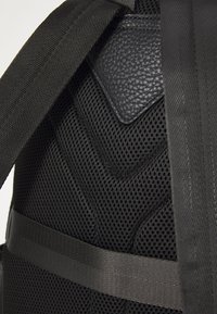 Sac à dos noir en matériau en maille avec un panneau en cuir texturé, des bretelles réglables et un motif en chevrons sur la surface.