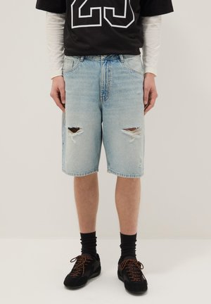 Persona che indossa pantaloncini di jeans strappati azzurri chiari, sneaker nere con lacci arancioni, calzini neri, maglia nera e camicia bianca a maniche lunghe.