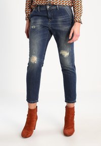 Mörkblå slitna jeans med trasiga knälappar, smal benfitting och uppvikta fållar. Matchas med orangea mockaskor med ankelskaft.