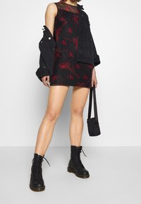 Black sleeveless dress with red CK logo and floral pattern, paired with a black denim jacket and black lace-up combat boots.