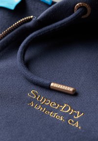 Marineblå hættetrøje med tekstureret stof. Har guld broderet "SuperDry Athletics, CA" og en bronzefarvet metalspids på snørebåndet.