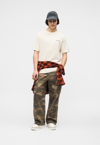 Νεαρός άνδρας που φοράει μπεζ μπλουζάκι McLaren, παντελόνι καμουφλάζ cargo, λευκά αθλητικά παπούτσια, μαύρο καπέλο και κόκκινο-μαύρο πουκάμισο καρό δεμένο γύρω από τη μέση του.