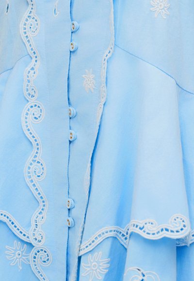 Ανοιχτό μπλε ύφασμα με λευκά κεντημένα λουλουδάτα και σπειροειδή σχέδια, με μικρά καλυμμένα με ύφασμα κουμπιά κατά μήκος της κεντρικής ραφής.