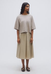 Pull court gris clair avec manches larges, associé à une jupe plissée beige. Le modèle porte des chaussures plates marron au design simple.