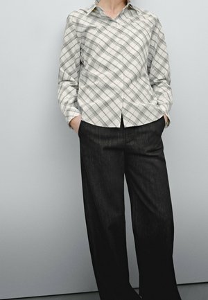 Personne portant une chemise à manches longues à carreaux blancs et gris, rentrée dans un pantalon large noir et ample, les mains dans les poches, sur un fond gris clair.