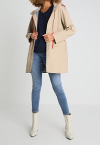 Beige voděodolná bunda s kapucí, nastavitelné oranžové šňůrky, dvě přední kapsy a šedé podšívání, spárovaná s modrými džíny a bílými kotníkovými botami.
