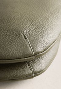 Gros plan sur un cuir grainé vert olive avec coutures visibles et bords pliés, montrant la texture et les détails du savoir-faire.