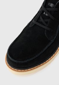 Black suede moccasin-style shoe with black laces and contrasting beige rubber sole showing detailed stitching.