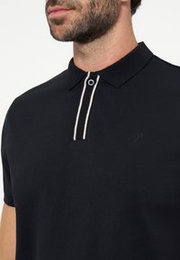 Homme portant un polo noir avec un col et une patte à rayures blanches, montrant le cou et le bas du visage avec une barbe.