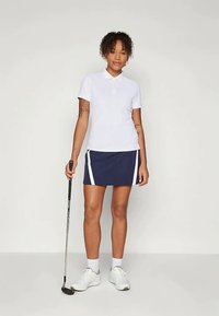 Λευκό πόλο μπλουζάκι με γιακά, συνδυασμένο με ναυτικό φούστα με λευκές πλευρικές ρίγες, φορεμένο με λευκά αθλητικά παπούτσια και κρατώντας μία γκολφ κλαρί.