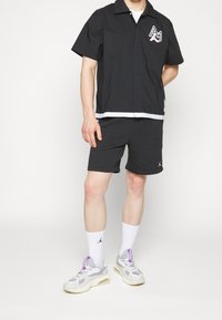 Svart kortärmad skjorta med krage och logotyp, tillsammans med svarta shorts. Bärs med vita strumpor och grå sneakers med lila detaljer.