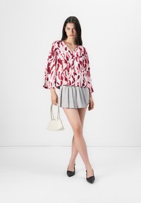 Lätt rosa blus med ett rött abstrakt mönster, ihopparad med en grå veckad kjol. Modellen håller en liten vit handväska och bär svarta klackar.