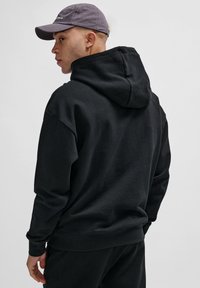Czarna bluza z kapturem wykonana z miękkiego materiału, o luźnym kroju i ściągaczach, w zestawie z szarym czapką. Widok z tyłu.
