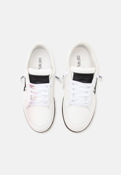 Ζευγάρι λευκά sneakers χαμηλού τύπου με λευκά κορδόνια, μαύρα ένθετα στη γλώσσα και κείμενο στην πάτο και στις πλαϊνές ετικέτες σε ανοιχτό φόντο.