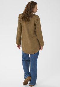 Blusa marrón de manga larga con tela texturizada y dobladillo redondeado, combinada con jeans azules rectos y zapatos marrones. Diseño minimalista.