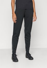 Pantaloni atletici negri cu picioare conice, confecționați dintr-un material neted. Prezintă accente reflectorizante și dungi laterale subtile pe partea inferioară a picioarelor.