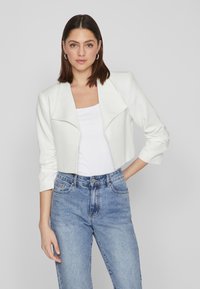 White cropped jacket with a wide collar and three-quarter sleeves, paired with a fitted white shirt and high-waisted blue jeans.