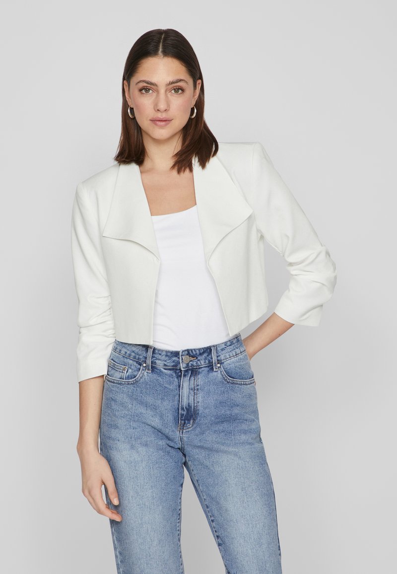White cropped jacket with a wide collar and three-quarter sleeves, paired with a fitted white shirt and high-waisted blue jeans.