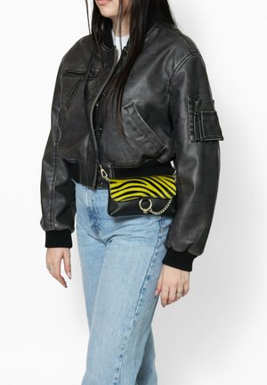 Giubbotto bomber in pelle nera con polsini a costine, abbinato a una borsa a tracolla gialla e nera a strisce con un accento a catena. Jeans azzurri chiari.