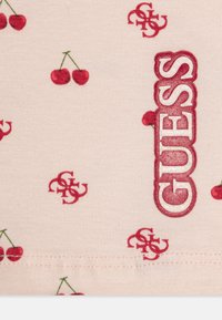 Material din bumbac roz, cu modele de cireșe roșii și un logo "GUESS" proeminent, realizat din litere texturate și lucioase.