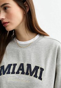 Felpa grigia con collo a giro, con stampa navy "MIAMI" e "Florida State" sottostante. Indossata sopra una camicia bianca, collana a catena sottile visibile.