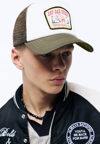 Weiße und olivgrüne Baseballmütze mit einem Front-Patch, das einen Bison zeigt, getragen mit einer schwarzen Satin-Bomberjacke und einer Perlenkette.