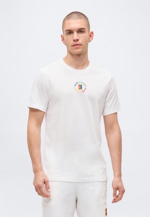 Mann mit kurzen Haaren trägt weißes Nike Court T-Shirt und Shorts, steht vor einem einfarbigen hellen Hintergrund.