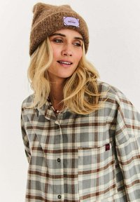 Kobieta z długimi blond włosami, nosząca brązową dzianinową czapkę i szarą koszulę w kratę z białymi i brązowymi wzorami, spoglądająca lekko na bok.