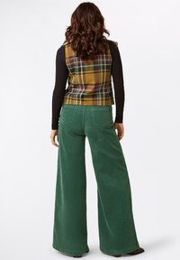 Karierte Weste in Braun und Gelb über einem schwarzen Langarmshirt; weite grüne Cordhosen; sichtbare Gesäßtaschen. Die Haare sind locker gewellt.