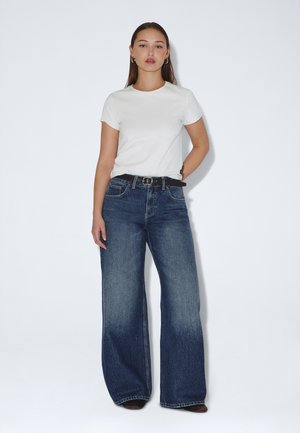 Donna in piedi contro un muro bianco, indossa una camicia bianca a maniche corte, jeans blu a gamba larga con cintura nera e orecchini a cerchio.