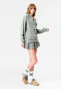 Cárdigan gris de gran tamaño y multi-textura con botones, combinado con una falda plisada. La modelo lleva calcetines blancos con patrones y zapatillas beige.