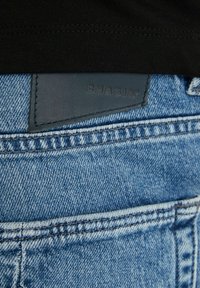 Mėlyni džinsai turi tekstūruotą paviršių, išryškintus siūlus ir juodą odinį ženklelį su užrašu "CHASIN'" gale.