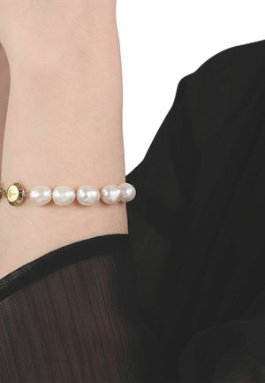 Polso adornato con un braccialetto di perle con chiusura rotonda in oro, mostrato su uno sfondo di manica nera trasparente.