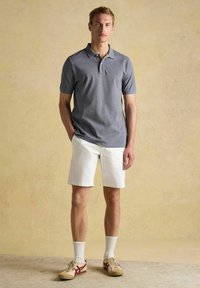 Sivý polo tričko s krátkymi rukávmi, biele šortky a béžové topánky s červenými akcentmi. Model stojí pred neutrálnym pozadím.