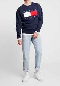 Felpa blu navy con design del logo bianco e rosso, collo rotondo, polsini a costine, abbinata a jeans azzurri risvoltati e sneakers bianche.