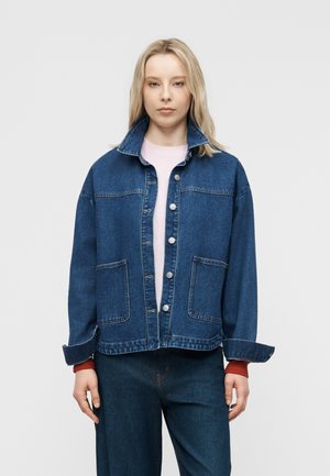 Blondynka w luźnej niebieskiej kurtce jeansowej z dużymi kieszeniami i guzikami, zestawiona z dopasowanymi dżinsami oraz jasnoróżową bluzką.