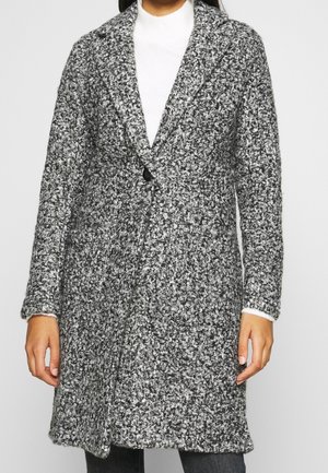 Vrouw draagt een halflange zwart-witte gebreide wollen jas over een witte coltrui, staat met ontspannen handen.