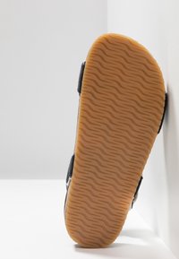 Sandal s texturovanou hnědou gumovou podrážkou, černým látkovým svrškem a nastavitelnými pásky. Podešev má vlnitý vzor pro lepší přilnavost.